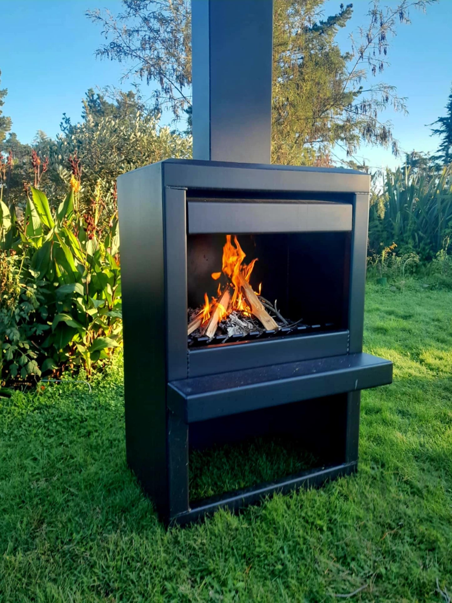 Outdoor steel firebox in corten finish, 800mm wide by 500mm deep, with a 1200mm high box and flue extending to 2470mm, set in a garden setting.