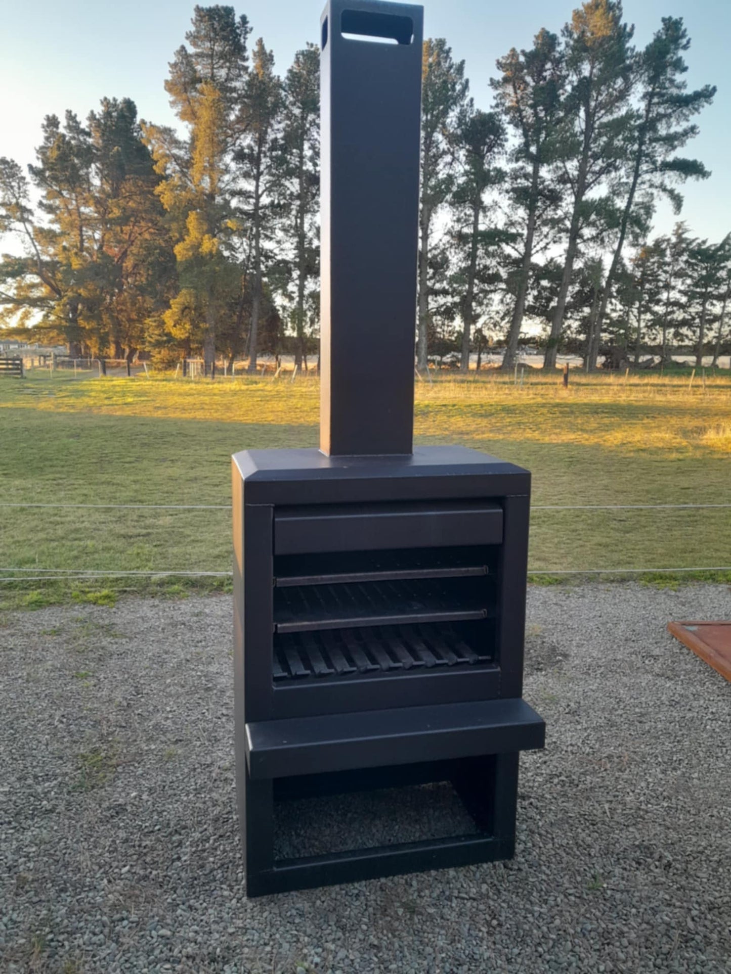 Outdoor steel firebox in corten finish, 800mm wide by 500mm deep, with a 1200mm high box and flue extending to 2470mm, set in a garden setting.
