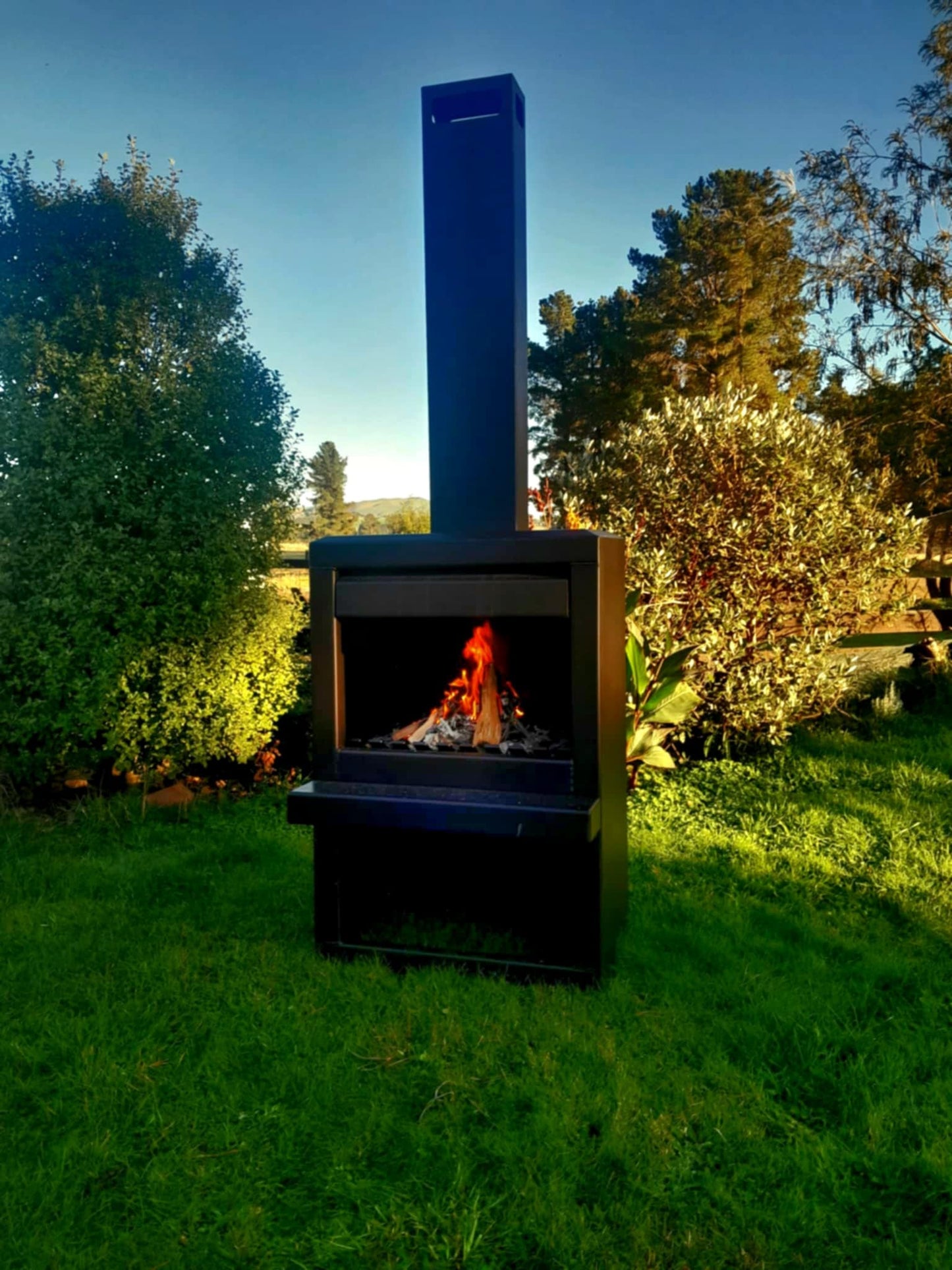 Outdoor steel firebox in corten finish, 800mm wide by 500mm deep, with a 1200mm high box and flue extending to 2470mm, set in a garden setting.
